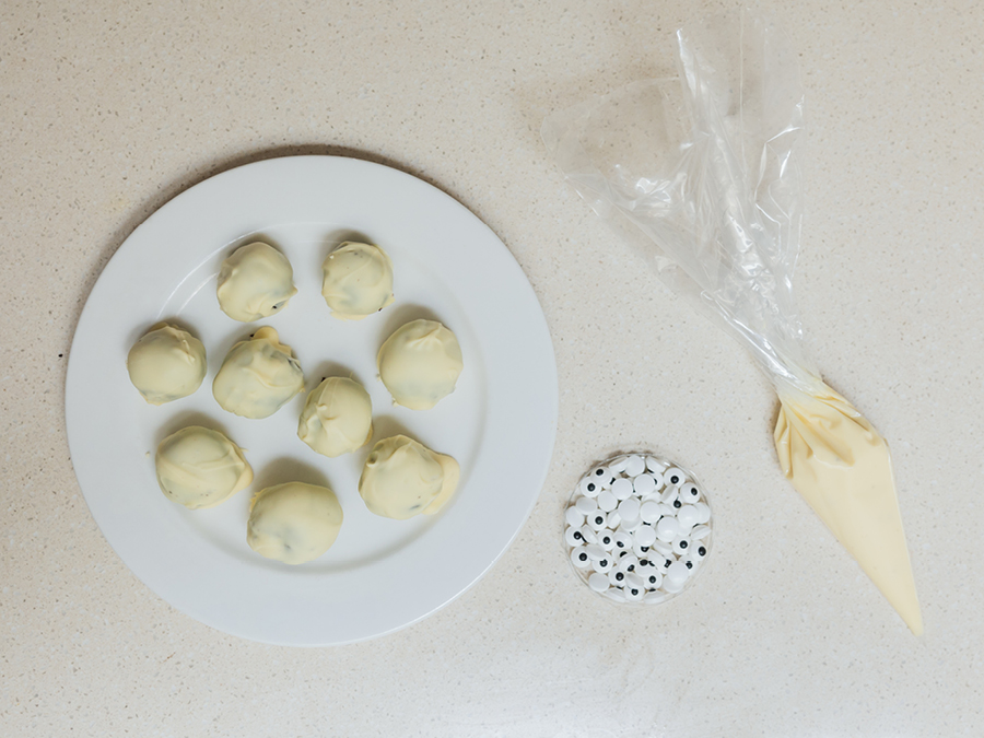 A white plate with rolled balls made from a mixture of Oreos and cream cheese, dipped in white chocolate. There is a bowl of candy eyes and a piping bag full of melted white chocolate.