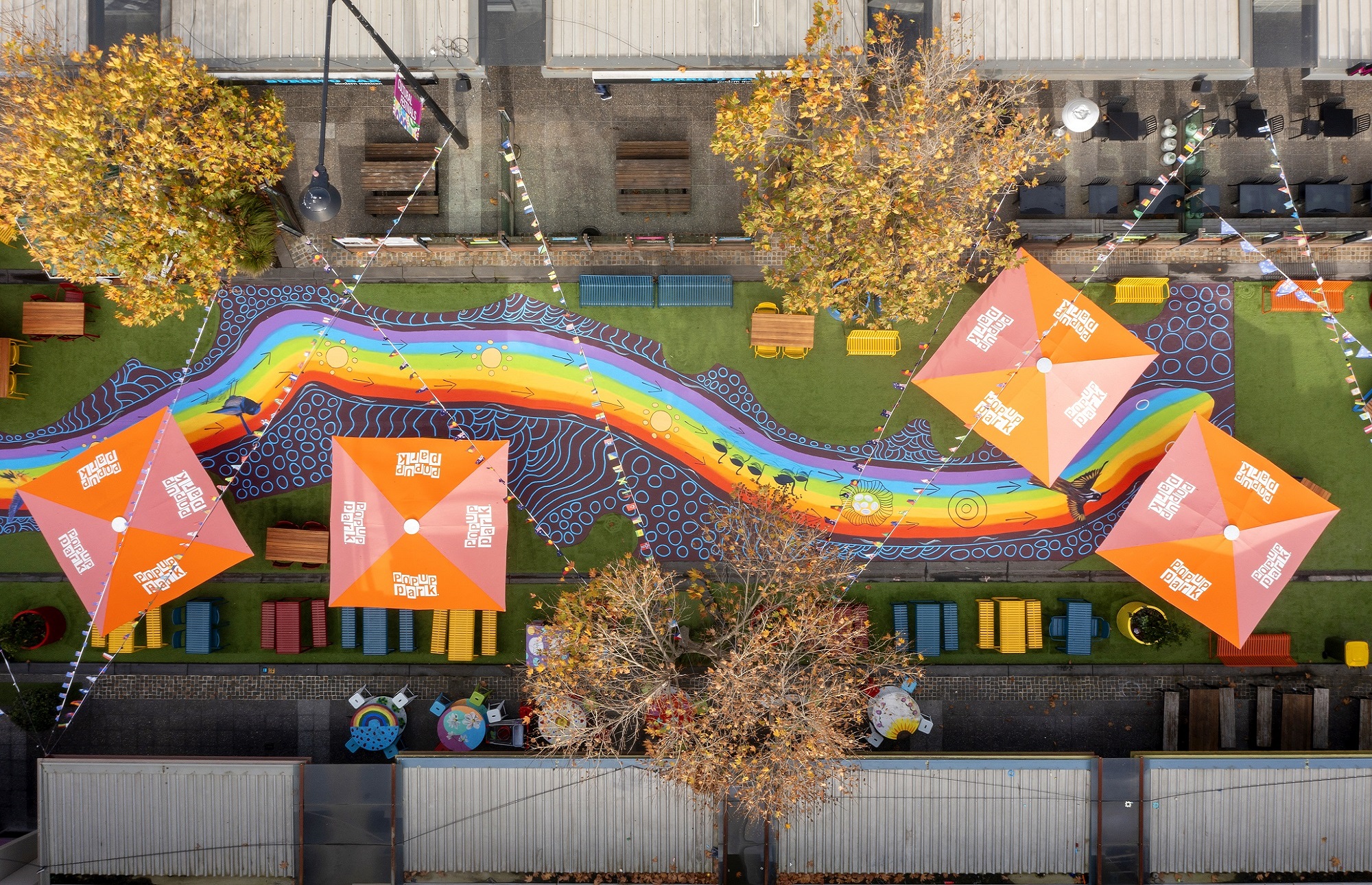 A picture taken by a drone directly above the Rainbow Serpent mural. The mural is on the ground in the middle of a green pop-up park that has orange umbrellas and colourful seating scattered around. People can walk on the mural and follow it as it winds directly through the middle of the park. It looks like a rainbow colored serpent. You can see some trees and the roofs of shops around the park.