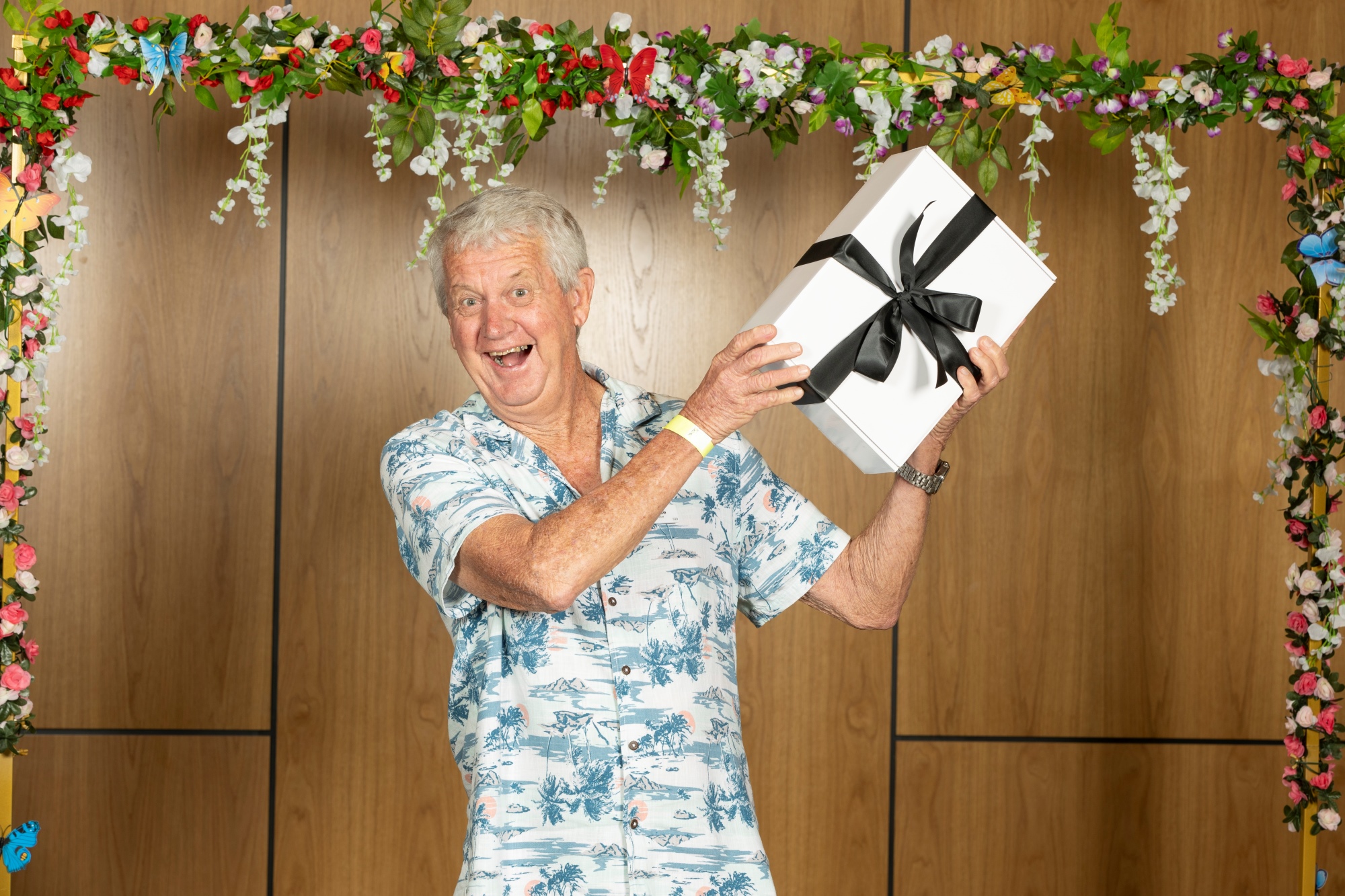 A smiling man holding a gift wrapped present in front of a flower arch