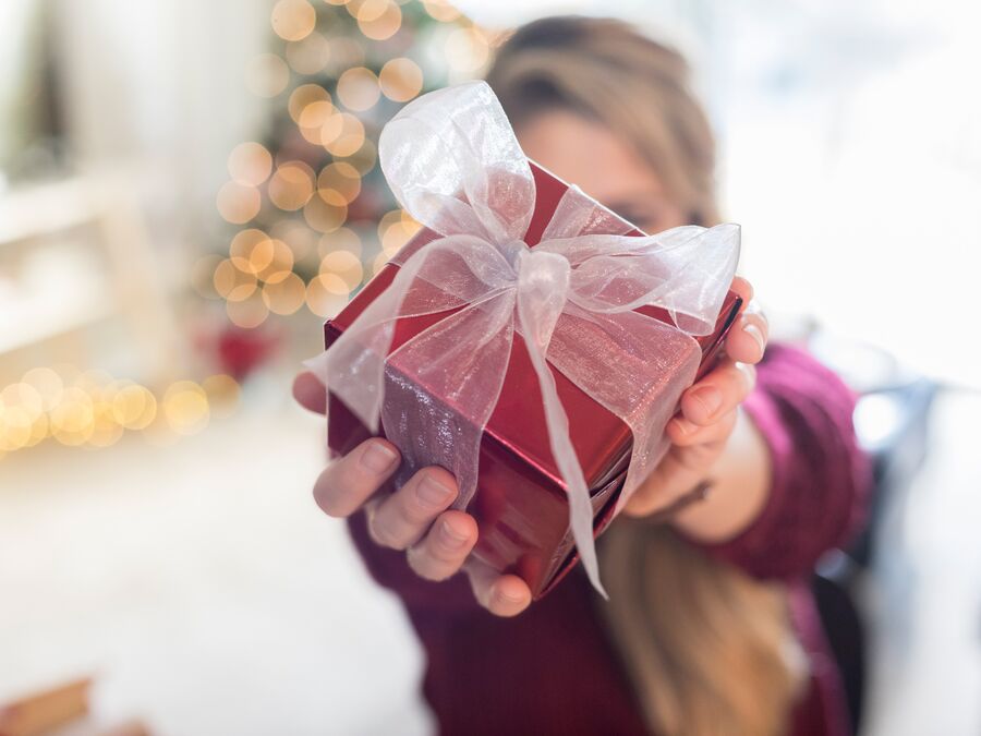 A red box is wrapped with a white sheer ribbon being held out in front of someone, covering their face. There is a blurred Christmas tree and living room in the background. 