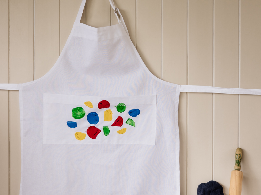 A white apron hanging on the wall decorated with colourful potato stamp design. 