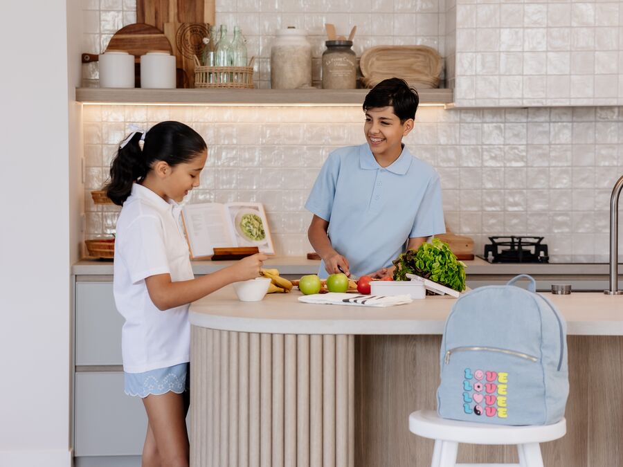 Two kids in a kitchen, one boy and one girl smiling as they chop up fruit to put in a lunchbox