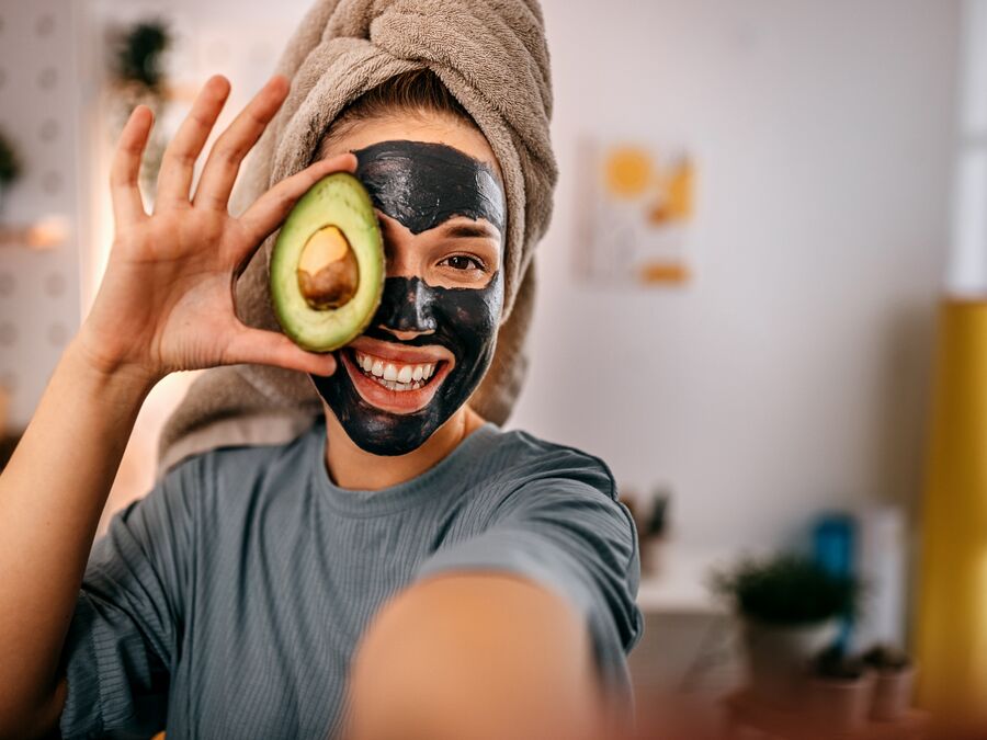 Young woman wearing a charcoal face mask and holding half a cut avocado in front of one eye.