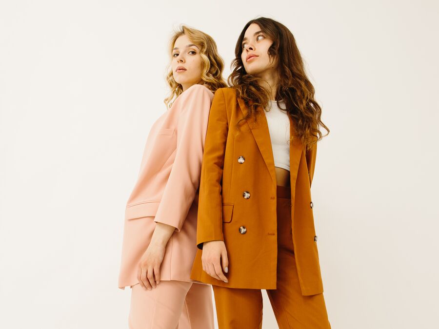 Two young females dressed in a colour-blocked blazer and pants. One is wearing a rust orange suit and the other is wearing a salmon coloured suit.