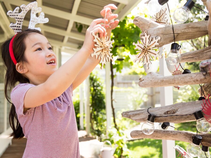 A little girl wearing reindeer antlers hangs a wooden snowflake decoration on a wooden-stick style Christmas tree outdoors