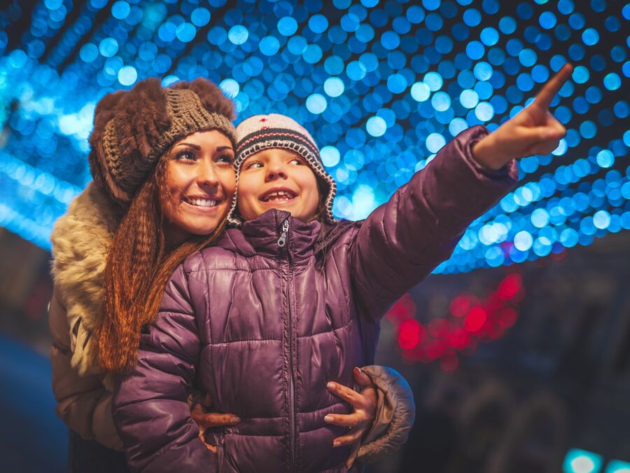 A mother and child stand under a net of blue fairly lights at night looking happy, with the kid pointing into the distance