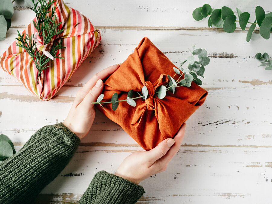 A gift wrapped in rust-coloured cloth with a sprig of eucalyptus leaves used as decoration, being held by two hands over a white timber background. A second cloth wrapped gift and leaves are next to it.